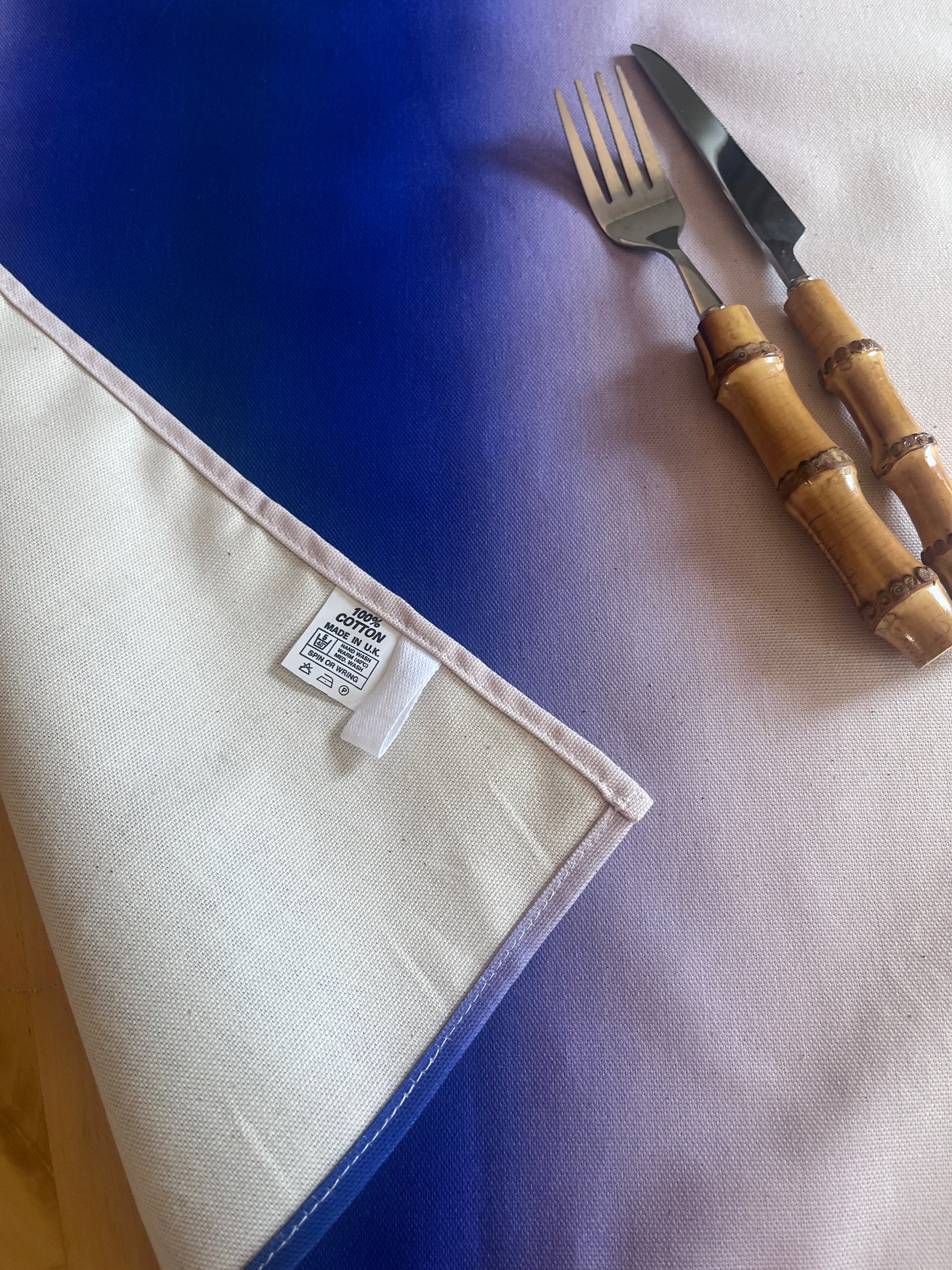 A square fabric napkin with a blue and white ombré gradient border, shown with a knife and fork on a matching gradient background. The napkin's reverse side is visible, showing a white woven tag.